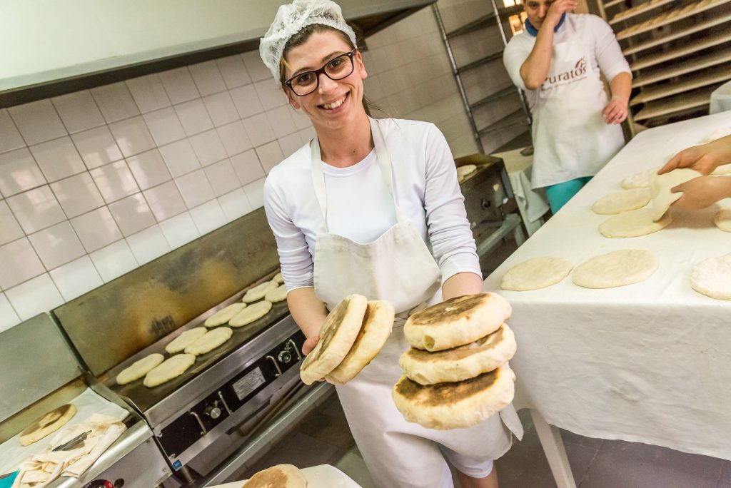 Die Küche mit allen Arbeitsschritten im Hintergrund und Frau mit frischen Brot in der Hand 