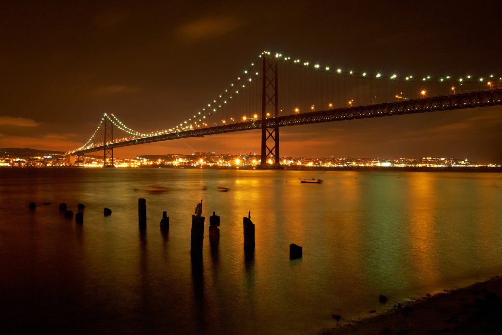 Brücke in nächtlicher Beleuchtung mit der leuchtenden Stadt im Hintergrund und dem Wasser im Vordergrund 