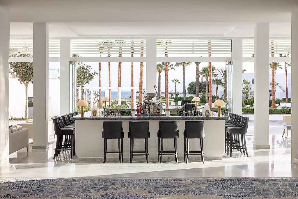 Grecotel Creta Palace, Stilvoll beleuchtete Hotelbar im Innenbereich mit Barhockern und einer großen Auswahl an Getränken.