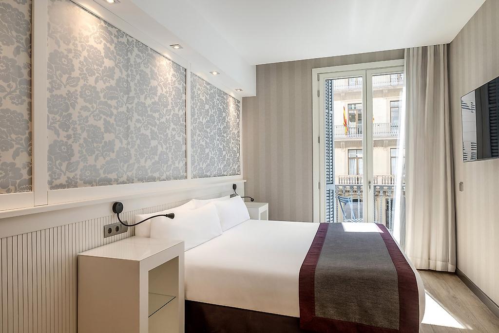 Catalonia Plaza Catalunya, Hell und modern eingerichtetes Doppelzimmer mit einem großen Doppelbett, stilvollem Holzkopfteil, Nachttischen, Designer-Lampen und einem großen Fenster, das viel Tageslicht hereinlässt.