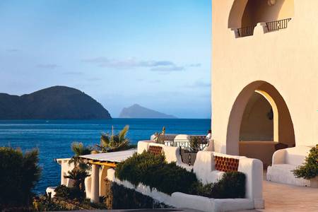 Therasia Resort Sea & Spa, Sonnige Außenterrasse mit Tischen und Stühlen vor einem Hotelgebäude.